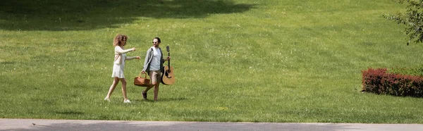 Full length of man carrying guitar and wicker basket near happy girlfriend gesturing in green park, banner — Foto stock