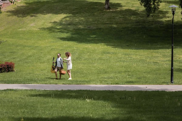 Full length of couple in summer clothes carrying guitar and picnic wicker basket in green park — Stock Photo