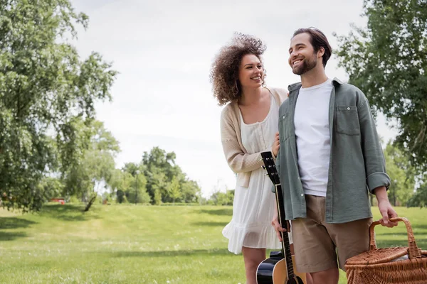 Man in summer clothes carrying guitar and wicker basket near happy girlfriend in green park — Foto stock