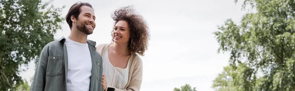 Happy curly woman looking at man in summer clothes in green park, banner — Stock Photo