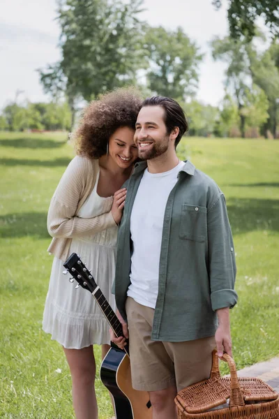 Man in summer clothes carrying guitar and wicker basket near cheerful girlfriend in green park — Stock Photo