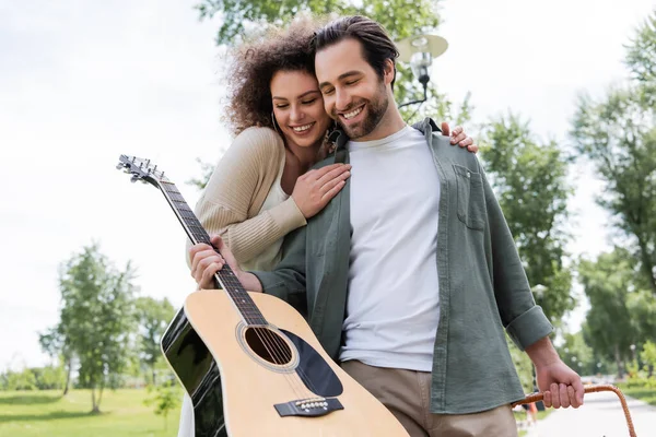 Homme souriant en vêtements d'été tenant la guitare près de petite amie heureuse dans le parc vert — Photo de stock