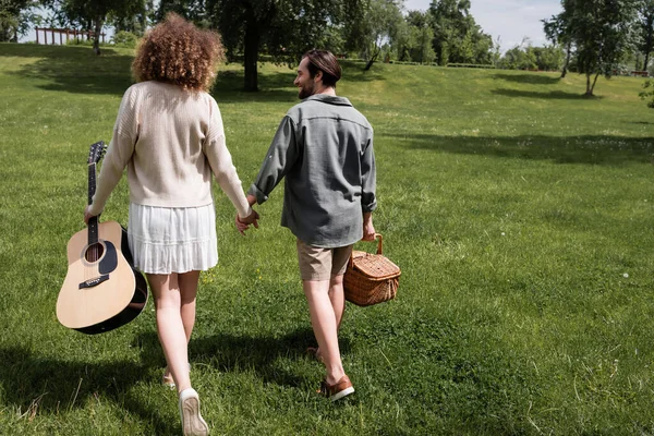 Back view of curly woman holding acoustic guitar while happy man carrying wicker basket in green park — Stockfoto