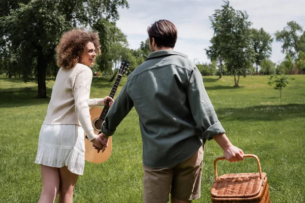 Happy curly woman holding acoustic guitar while looking at man carrying wicker basket in green park — Fotografia de Stock