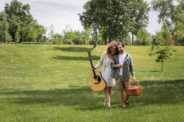 Comprimento total de mulher encaracolado feliz segurando guitarra acústica perto do homem carregando cesta de vime enquanto caminhava no parque verde — Fotografia de Stock
