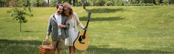 Happy curly woman holding acoustic guitar near man with wicker basket and walking in green park, banner — Stock Photo