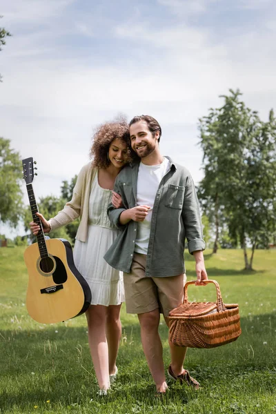 Mujer rizada feliz sosteniendo la guitarra acústica cerca de novio con canasta de mimbre en el parque verde - foto de stock