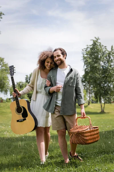 Happy curly woman holding acoustic guitar near boyfriend with wicker basket in green park — Stockfoto