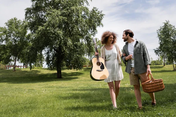 Full length of smiling curly woman holding acoustic guitar near boyfriend with wicker basket walking in park — Fotografia de Stock