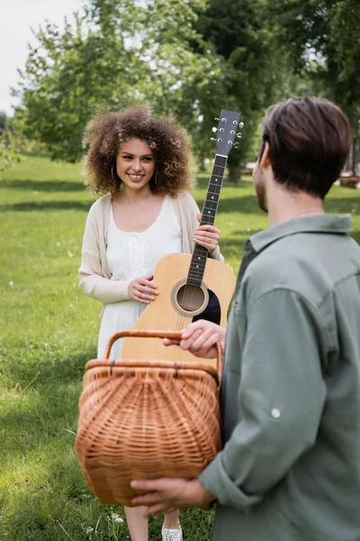 Glücklich lockige Frau mit Akustikgitarre und Blick auf Freund mit Weidenkorb im grünen Park — Stockfoto