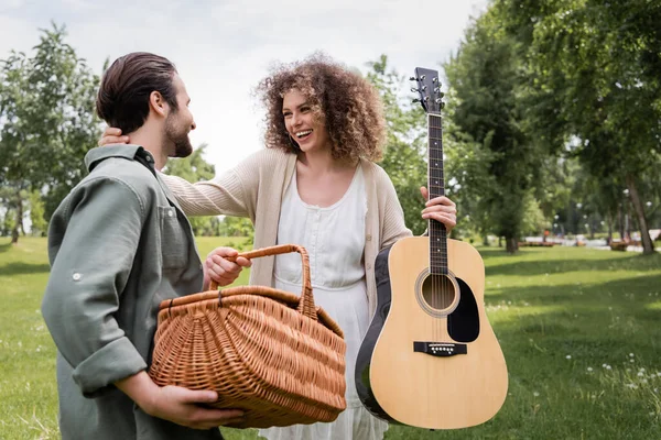 Happy curly woman holding acoustic guitar and hugging boyfriend with wicker basket in green park - foto de stock