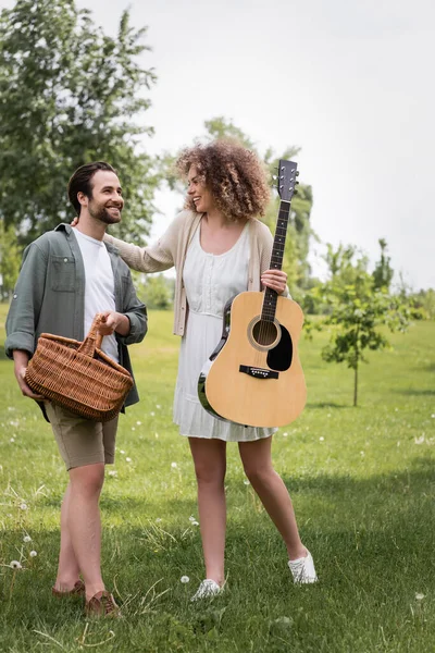 Longitud completa de la mujer rizada sosteniendo la guitarra acústica cerca novio feliz con cesta de mimbre en el parque verde - foto de stock