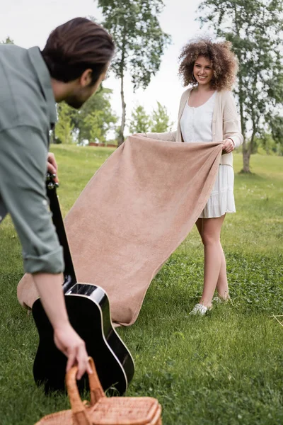 Cheerful curly woman in summer clothes holding blanket near boyfriend with acoustic guitar in park — Stockfoto