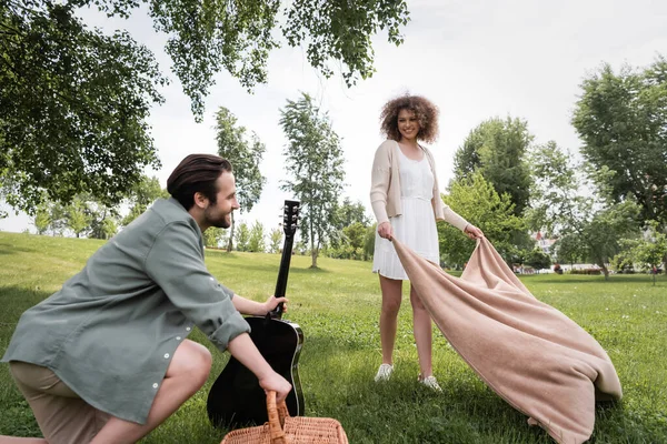 Pleased curly woman in summer clothes holding blanket near boyfriend in park — Stock Photo