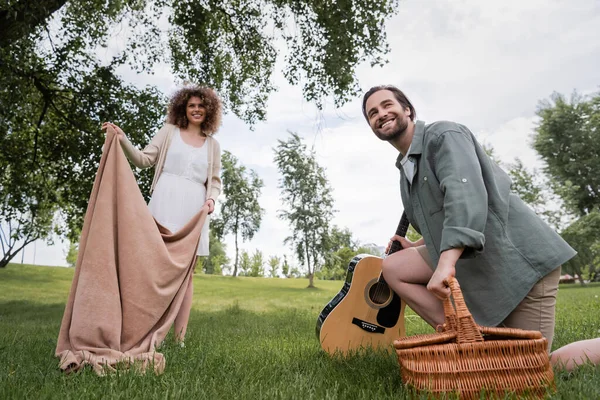 Low angle view of cheerful woman in summer clothes holding blanket near boyfriend with wicker basket in park — Stock Photo