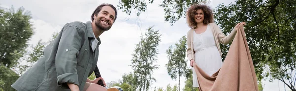 Low angle view of cheerful curly woman in summer clothes holding blanket near boyfriend in park, banner - foto de stock