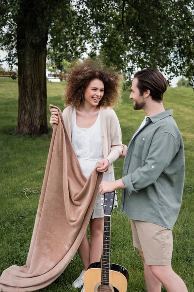 Весела кучерява жінка в літньому одязі тримає ковдру біля хлопця в парку — стокове фото