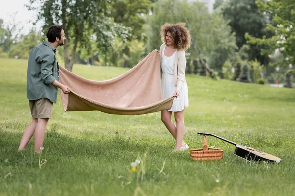 Joyful young couple in summer clothes putting picnic blanket on green grass in park — Photo de stock