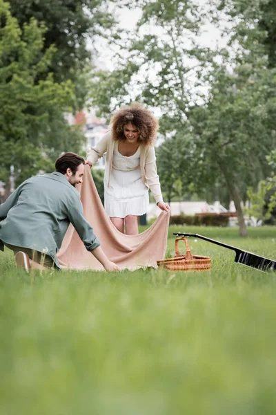 Happy young couple in summer clothes putting picnic blanket on green grass in park — Stock Photo