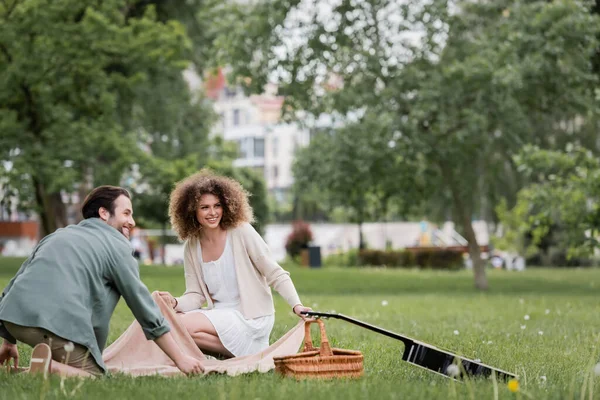 Happy couple sitting on blanket near picnic wicker basket and acoustic guitar — Stockfoto