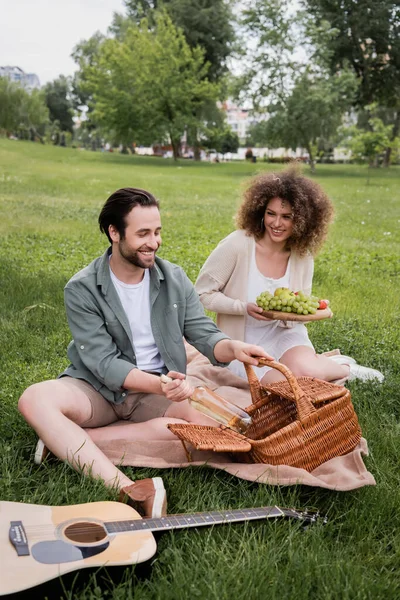 Happy man taking bottle from wicker basket near curly girlfriend holding cutting board with fruits during picnic — Foto stock