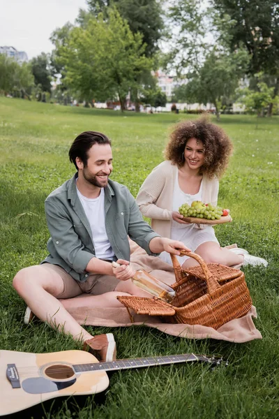 Happy man taking bottle from wicker basket near curly girlfriend holding cutting board with fruits during picnic — Stock Photo