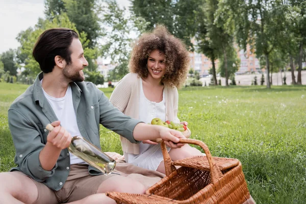 Cheerful man taking bottle from wicker basket near curly woman holding cutting board with fruits during picnic — Foto stock