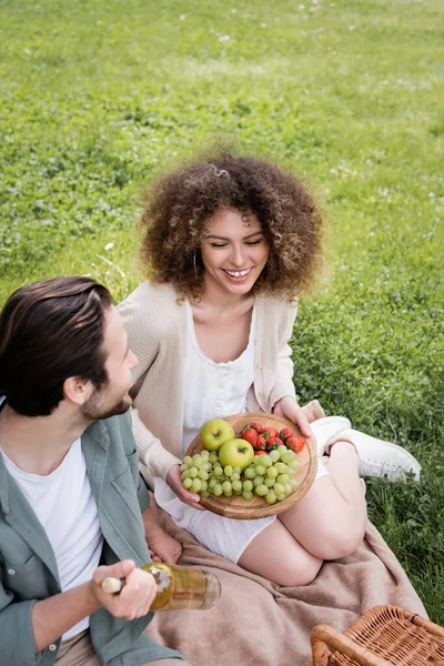 High angle view of happy man holding bottle of wine near curly girlfriend sitting with cutting board and fruits — Stockfoto