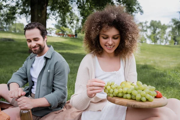 Mulher encaracolado segurando placa de corte com frutas frescas perto de namorado feliz abertura garrafa de vinho — Fotografia de Stock