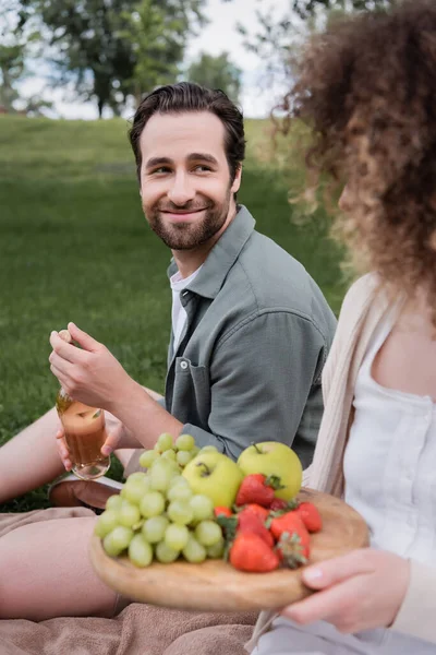 Happy man opening bottle of wine near curly girlfriend sitting with fruits during picnic — Fotografia de Stock