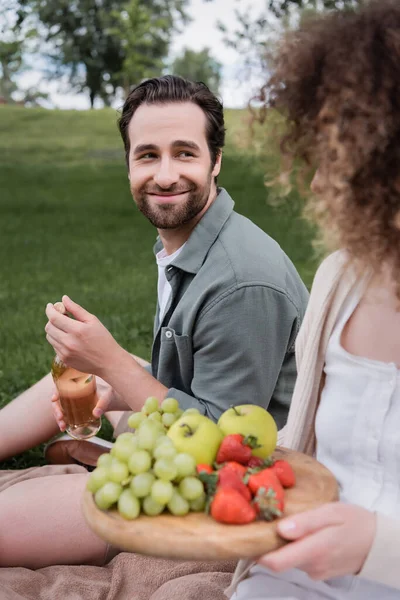 Happy man opening bottle of wine near curly girlfriend sitting with fruits during picnic — Stock Photo