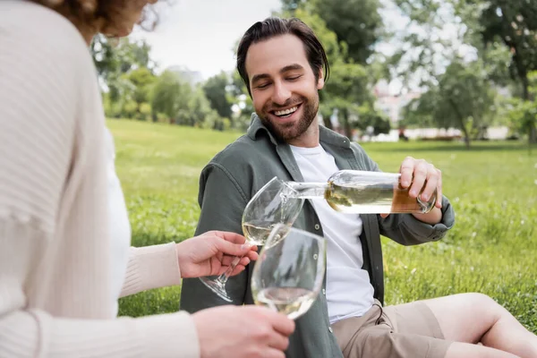 Homem barbudo feliz derramando vinho em vidro perto da namorada durante o piquenique — Fotografia de Stock