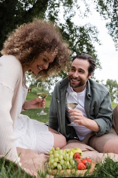 Happy man looking at cheerful curly woman with glass of wine during summer picnic — Stock Photo