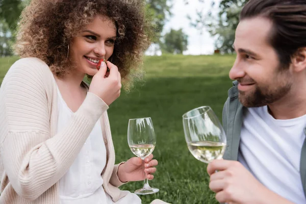 Feliz rizado mujer con vaso de vino comer fresa fresca cerca de novio - foto de stock