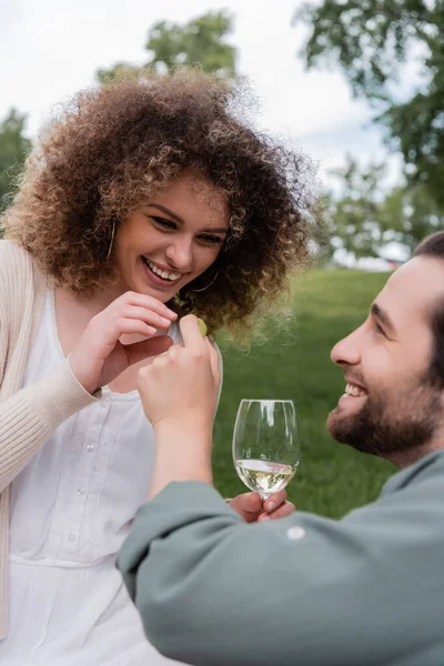 Happy man with glass of wine holding fresh grape near cheerful girlfriend during picnic — Photo de stock