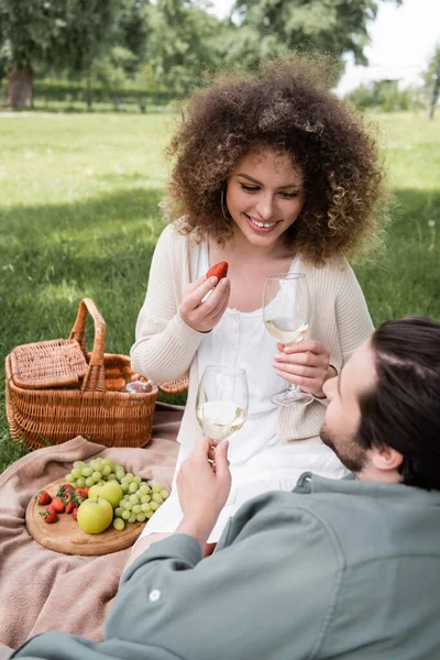 Glücklich lockige Frau hält Erdbeere und ein Glas Wein neben ihrem lächelnden Freund beim Picknick — Stockfoto