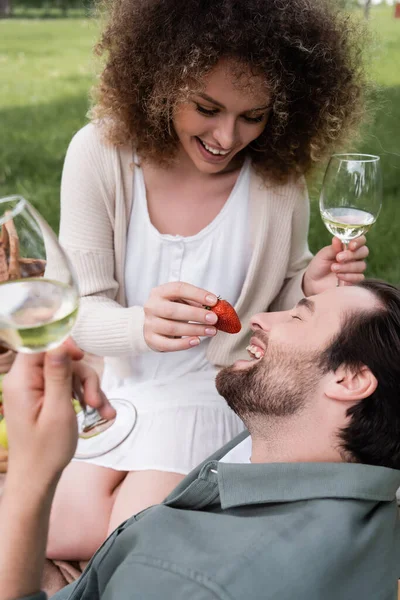 Happy curly woman holding glass of wine and feeding boyfriend with fresh strawberry during picnic — Stock Photo