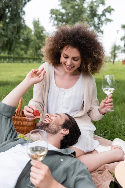 Cheerful curly woman holding glass of wine and feeding boyfriend with fresh strawberry during picnic — Fotografia de Stock