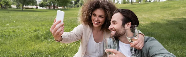 Счастливая молодая пара делает селфи на смартфоне во время летнего пикника в парке, баннер — стоковое фото