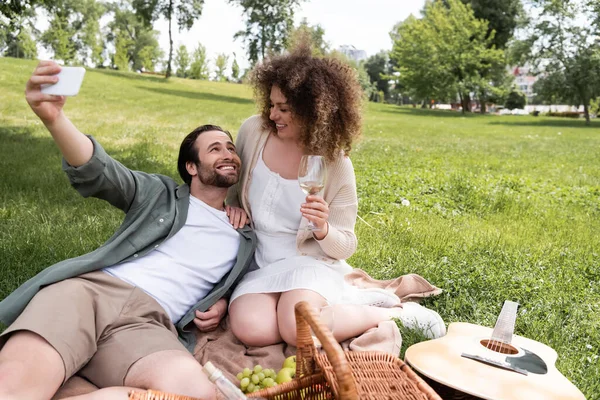 Joyful young couple taking selfie on smartphone during summer picnic in park — Stockfoto