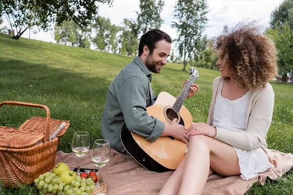 Hombre feliz tocando la guitarra acústica cerca de la mujer rizada durante el picnic de verano - foto de stock