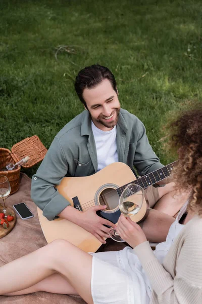 Hochwinkelaufnahme eines romantischen Mannes beim Spielen einer akustischen Gitarre in der Nähe einer lockigen Frau mit einem Glas Wein beim Picknick — Stockfoto