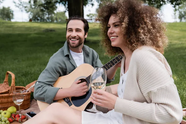 Cheerful man playing acoustic guitar near happy woman with glass of wine during picnic — Foto stock