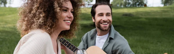 Романтичный мужчина, играющий на акустической гитаре рядом с кудрявой женщиной во время пикника, баннер — стоковое фото