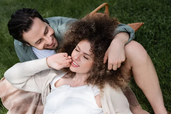 Draufsicht der lockigen Frau, die beim Picknick im Sommerpark auf ihrem glücklichen Freund liegt — Stockfoto
