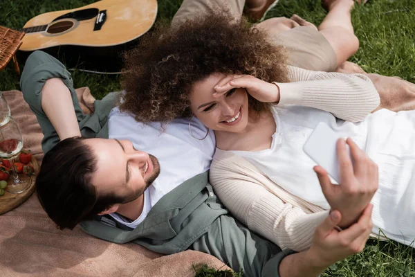 Vue de dessus de femme heureuse prenant selfie sur smartphone tout en étant couché avec petit ami pendant le pique-nique — Photo de stock