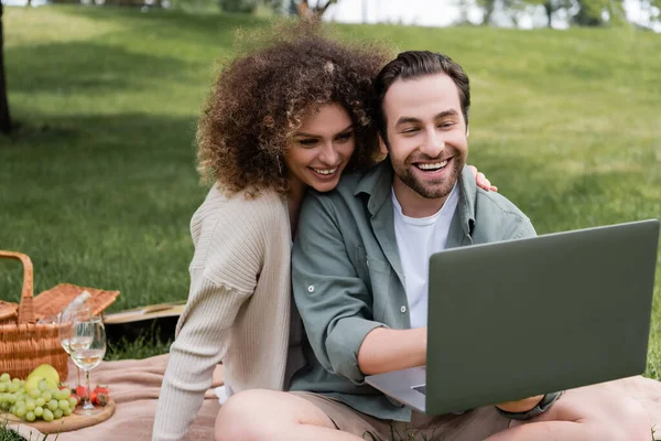 Happy man using laptop near curly girlfriend during picnic in park - foto de stock