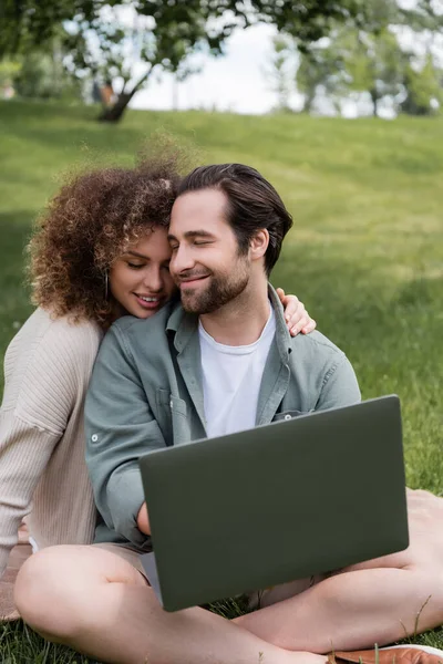 Кучерява жінка посміхається, обіймаючи хлопця з ноутбуком, що працює дистанційно в парку — стокове фото