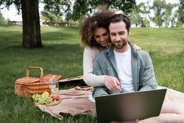 Mujer rizada apuntando a la computadora portátil cerca de novio durante el picnic en el parque - foto de stock