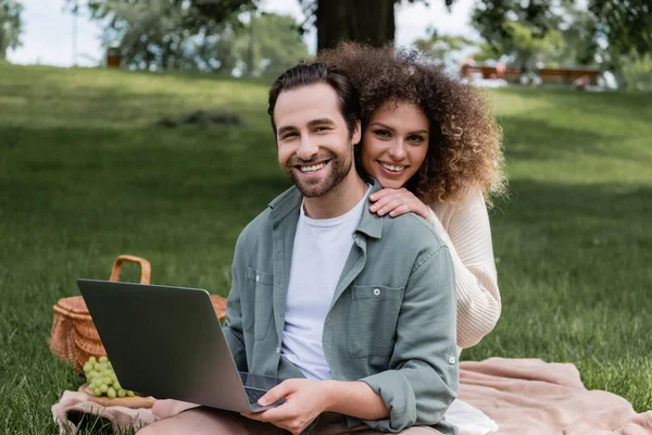 Cheerful man holding laptop and smiling with curly woman during picnic — Fotografia de Stock